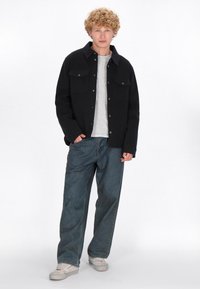 Giacca nera con bottoni e due tasche sul petto sopra una maglietta grigia chiara, abbinata a dei jeans blu in denim larghi e sneakers bianche.