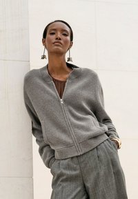 Grauer Reißverschluss-Pullover mit strukturierter Oberfläche, gerippten Bündchen und Kragen, kombiniert mit gestreiften Hosen und dekorativen Ohrringen.