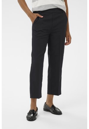 Person trägt knielange schwarze Hose, eine Hand in der Tasche, kombiniert mit schwarzen Leder-Loafers und einem grau-weiß gestreiften Oberteil.