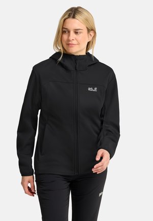 Blonde vrouw met een zwarte Jack Wolfskin jas en broek, staand tegen een effen lichte achtergrond, lichtjes opzij kijkend.