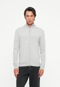 Man in een lichtgrijze zip-up sweater en zwarte broek, staand tegen een vlakke witte achtergrond, met het gezicht naar voren.
