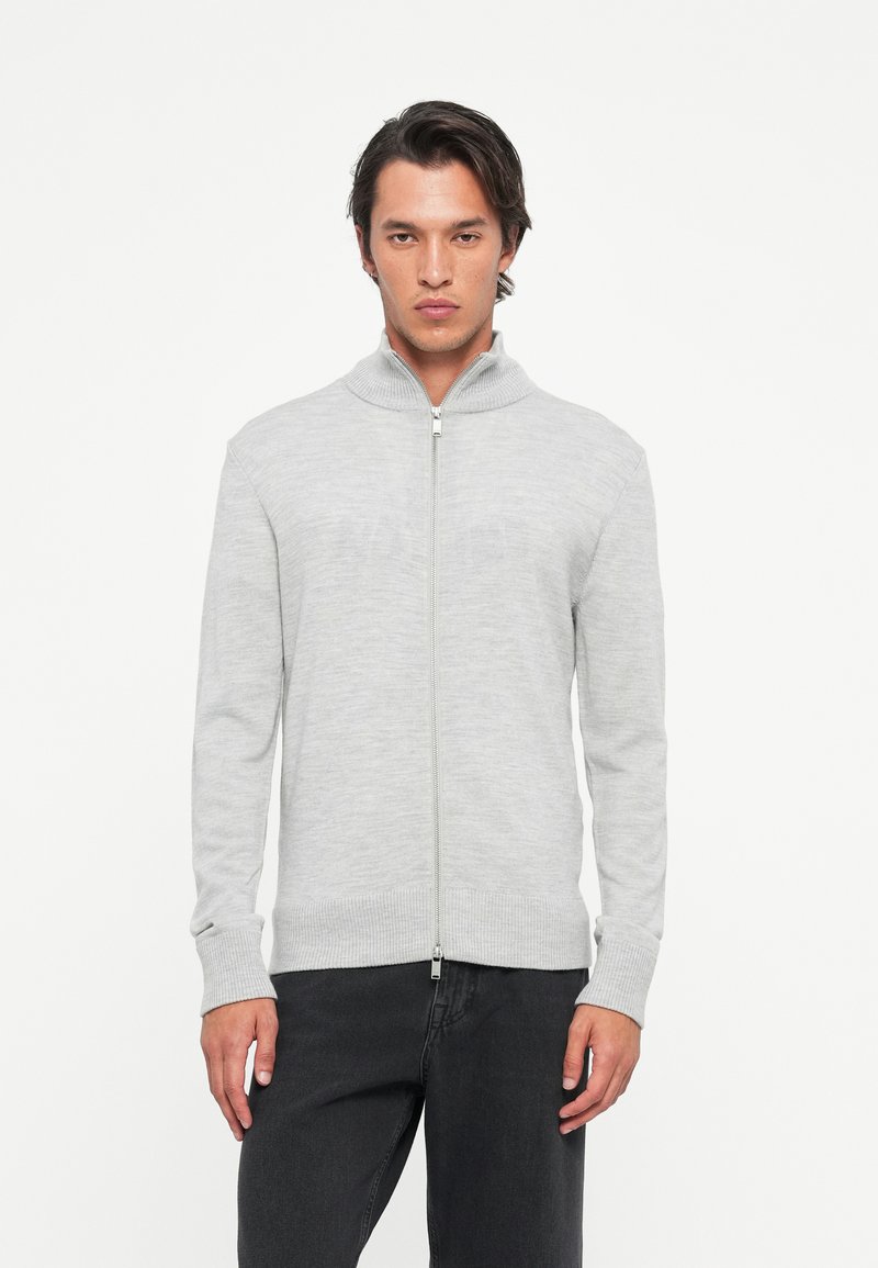 Man in een lichtgrijze zip-up sweater en zwarte broek, staand tegen een vlakke witte achtergrond, met het gezicht naar voren.