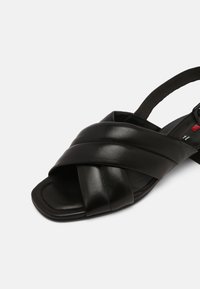 Sandale en cuir noir avec des brides croisées, un bout ouvert et une semelle plate. Dispose d'une sangle arrière pour le soutien et un design minimaliste.