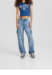 Cropped blue T-shirt with white text paired with baggy light blue cargo jeans featuring side pockets and black sneakers with white accents.