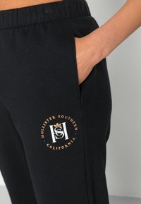 Svarta bomullsjoggers med sidoficka, med en rund logotyp i vitt och guld som säger "Hollister Southern California."
