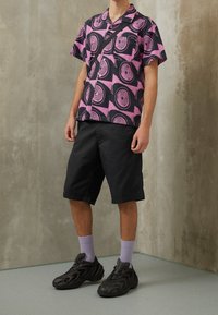 Lyhythihainen paita, jossa on vaaleanpunainen ja musta spiraalikuvio, yhdistettynä mustiin shortseihin ja teksturoituihin mustiin lenkkareihin, joissa on harmaat sukat.