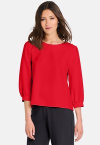 Blusa rossa a maniche lunghe con scollo tondo, tessuto morbido e maniche leggermente a palloncino, abbinata a pantaloni scuri.