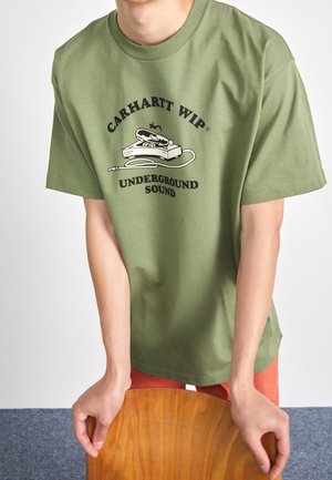 Person trägt ein olivgrünes Carhartt WIP T-Shirt mit einer Grafik eines Plattenspielers und dem Text „Underground Sound“, Hände ruhen auf einem Holzstuhl.