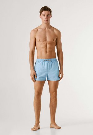 Noor mees seisab paljajalu, kandes helesiniseid ujumispükse, valge ühtlase taustaga.