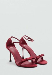 Sandali con tacco alto in raso rosso, dotati di cinturini alla caviglia e caratterizzati da un elegante fiocco frontale e una silhouette slanciata. Texture liscia e finitura lucida.