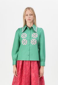 Camisa verde de botones con cuello puntiagudo, que presenta bordados de flores rosas en el frente y mangas fruncidas. Textura de tela suave.