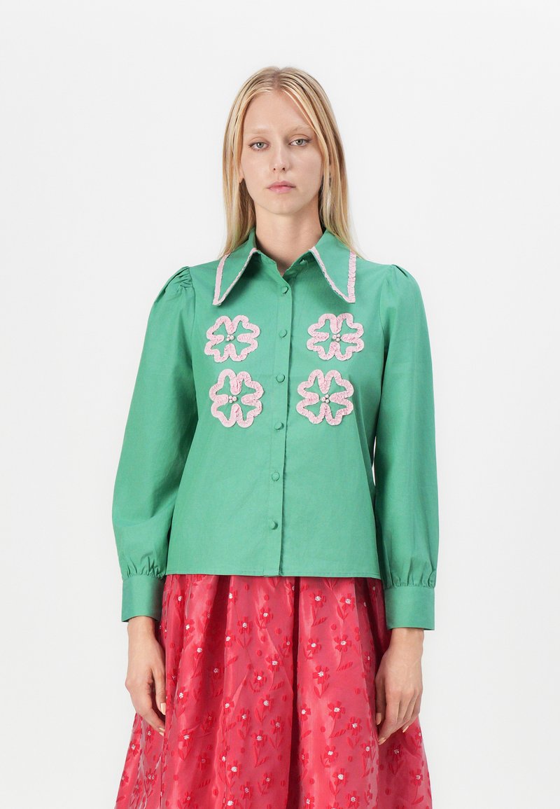 Camisa verde de botones con cuello puntiagudo, que presenta bordados de flores rosas en el frente y mangas fruncidas. Textura de tela suave.