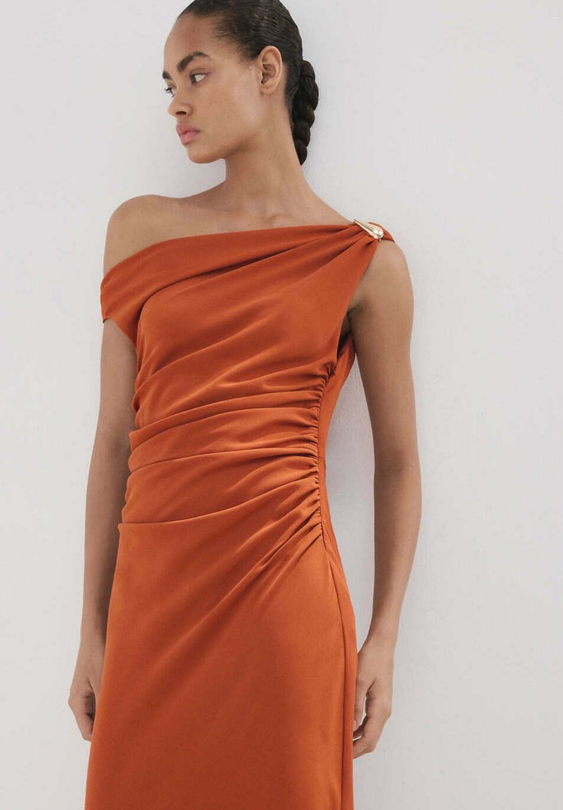 Femme aux cheveux tressés portant une robe rouille froncée asymétrique avec un détail doré à l'épaule, debout devant un mur clair uni.