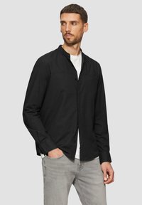 Chemise noire à manches longues avec un col à boutons, une poche poitrine unique et une texture lisse, associée à un t-shirt blanc et un jean gris.