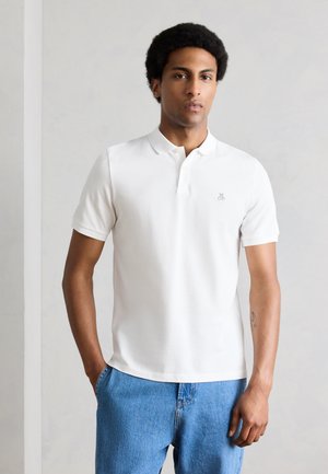 Άνδρας στέκεται σε απλή τοίχο φορώντας λευκό κοντομάνικο πουκάμισο πόλο και μπλε τζιν, με τα χέρια στις τσέπες, με ορατό τατουάζ στο αριστερό αντιβράχιο.