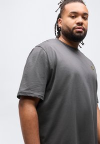 Άνδρας με πλεξούδες στα μαλλιά και γενειάδα, φορώντας σκοτεινό γκρι μπλουζάκι με μικρό κίτρινο λογότυπο στο στήθος, μπροστά σε απλό φωτεινό φόντο.