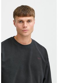 Svart genser med rund hals, ribbet kant nederst og på ermene. Liten logo i kastanjebrun på brystet. Glatt tekstur og avslappet passform.