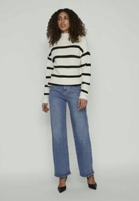 Balts un melns svītrots džemperis ar augstu apkakli, saskaņots ar gaiši ziliem plašiem biksēm un melniem norādītiem apaviem.