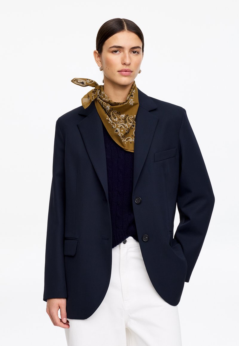 Femme aux cheveux foncés attachés, portant une écharpe à motif doré, un blazer bleu marine, un haut noir et un pantalon blanc, debout devant un fond uni.