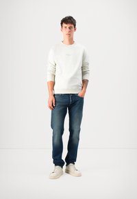 Young man standing with one hand in pocket, wearing a white sweatshirt, blue jeans, and white sneakers against a plain white background.