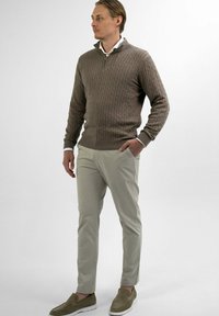 Bruin kabelgebreide trui met een kwart rits, gecombineerd met lichtbeige chino's en groene instappers. Neutrale achtergrond.