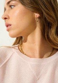 Guldörhängen i hoop-stil och ett lager av guldhalsband som bärs med en ljusrosa texturerad tröja med ribbat nackband.