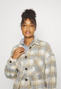 Casaco de fleece xadrez com tons de cinza, creme e amarelo, com fechos de botão e cintura com cinto. Textura suave e design oversized.