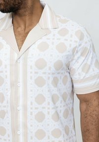 Chemise à manches courtes boutonnée de couleur beige clair avec un motif géométrique blanc. Présente un col en V et une texture de tissu lisse.