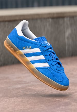 Blauwe Adidas Gazelle sneaker met witte strepen en gumzool, getoond midden in de lucht tegen een wazige betonnen achtergrond.
