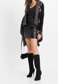 Cardigan nero a maglia con motivo geometrico, abbinato a una gonna mini in ecopelle nera con bottoni frontali e stivali neri in suede con tacco alti fino al ginocchio.