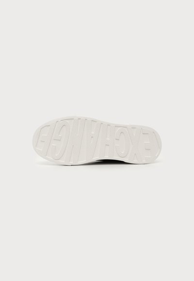 Semelle d'une chaussure blanche avec un texte en gras et en relief épelant "EXCHANGE", visible en dessous sur un fond clair.