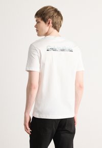 Balts kokvilnas T-krekls ar īsām piedurknēm, aizmugurē ar sudraba Calvin Klein logo un klasisku apaļu kakla dizainu. Redzami melni bikses.
