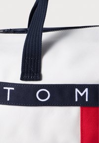 Τσάντα από καμβά με ναυτικοδέ χερούλια, λευκό σώμα κα ι κόκκινη λεπτομέρεια. Η λέξη "TOM" εμφανίζεται με λευκά γράμματα σε μια ναυτική λουρίδα. Υφασμάτινη υφή.