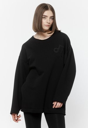 Młoda kobieta o prostych brązowych włosach nosi oversize'ową czarną bluzę z subtelnym haftowanym motywem nuty na piersi na białym tle.