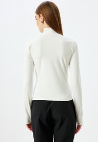 Wit, lang mouwtop met een hoge kraag, een gladde textuur en een aansluitend ontwerp, gedragen met zwarte broek, afgebeeld van achteren.
