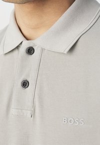 Gros plan sur le col d'une chemise polo gris pâle avec deux boutons noirs et le logo "BOSS" brodé sur la poitrine.
