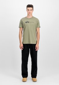 Olivza zaļa kokvilnas t-krekls ar melnu "ALPHA INDUSTRIES" logo, saskaņots ar melnām biksēm un brūniem zābakiem. Vienkāršs apļa kakla dizains.