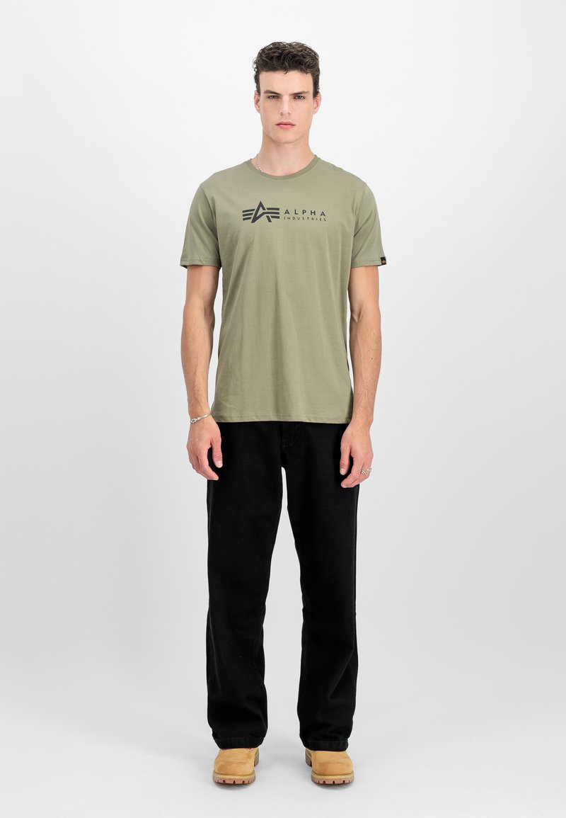 Olivza zaļa kokvilnas t-krekls ar melnu "ALPHA INDUSTRIES" logo, saskaņots ar melnām biksēm un brūniem zābakiem. Vienkāršs apļa kakla dizains.