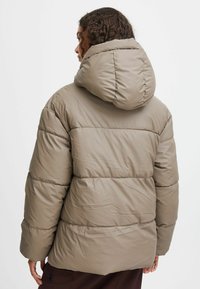 Pufferjakke i lys brun, med hætte, quiltet design og afslappet pasform. Materialet fremstår glat og let skinnende.