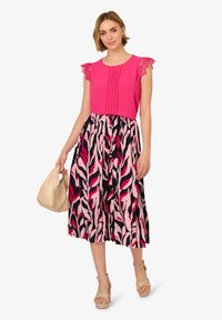 Top rosa con accenti in pizzo, dettagli plissettati e una gonna a fantasia floreale nei toni del rosa, rosso e blu navy. Scarpe e borsa beige completano l'outfit.