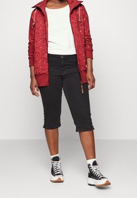 Person iført en rød mønstret hættetrøje med lynlås, hvid skjorte, sorte knælange bukser og sorte sneakers med høj top, hvide såler og snørebånd.