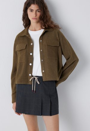 Frau trägt eine braune Jacke mit Knöpfen über einem weißen Hemd und einem dunkel plissierten Rock, der an der Taille gebunden ist, stehend vor einer schlichten Wand.