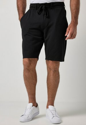 Homme portant un short noir à cordon et des baskets blanches, debout les mains détendues le long du corps sur un fond clair uni.
