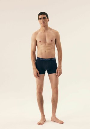 Black boxer briefs with a blue "HENDERSON" logo on the waistband. The model has a slim build and is barefoot. Neutral background.
