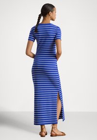 Žena s pletenými vlasmi oblečená v modro-bielych pruhovaných maxi šatách s krátkym rukávom a bočným rozparkom, v hnedej plochej obuvi, stojaca pred jednofarebným pozadím.