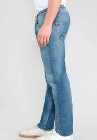 Jean en denim bleu clair avec une coupe droite, comportant cinq poches et des coutures contrastées. Porté avec des baskets blanches et une chemise unie.