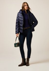 Chaqueta acolchada azul marino con capucha, cierre de cremallera, combinada con un suéter azul marino, jeans oscuros, botas de tobillo marrones y un bolso pequeño verde.