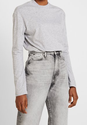 Osoba ubrana w jasnoszarą koszulę z długim rękawem, wciągniętą do wysokich, spranych szarych jeansów z przednimi kieszeniami i szlufkami na pasek.