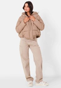 Giacca a vento beige con cappuccio, design trapuntato ed orlo elasticizzato. Abbinata a pantaloni cargo beige e scarpe bianche con strisce.