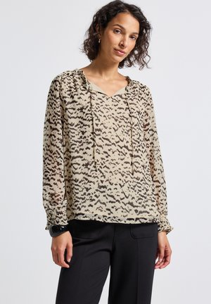 Vrouw met krullend haar draagt een beige blouse met patroon en een koordhals, samen met zwarte broek, staande tegen een effen witte achtergrond.